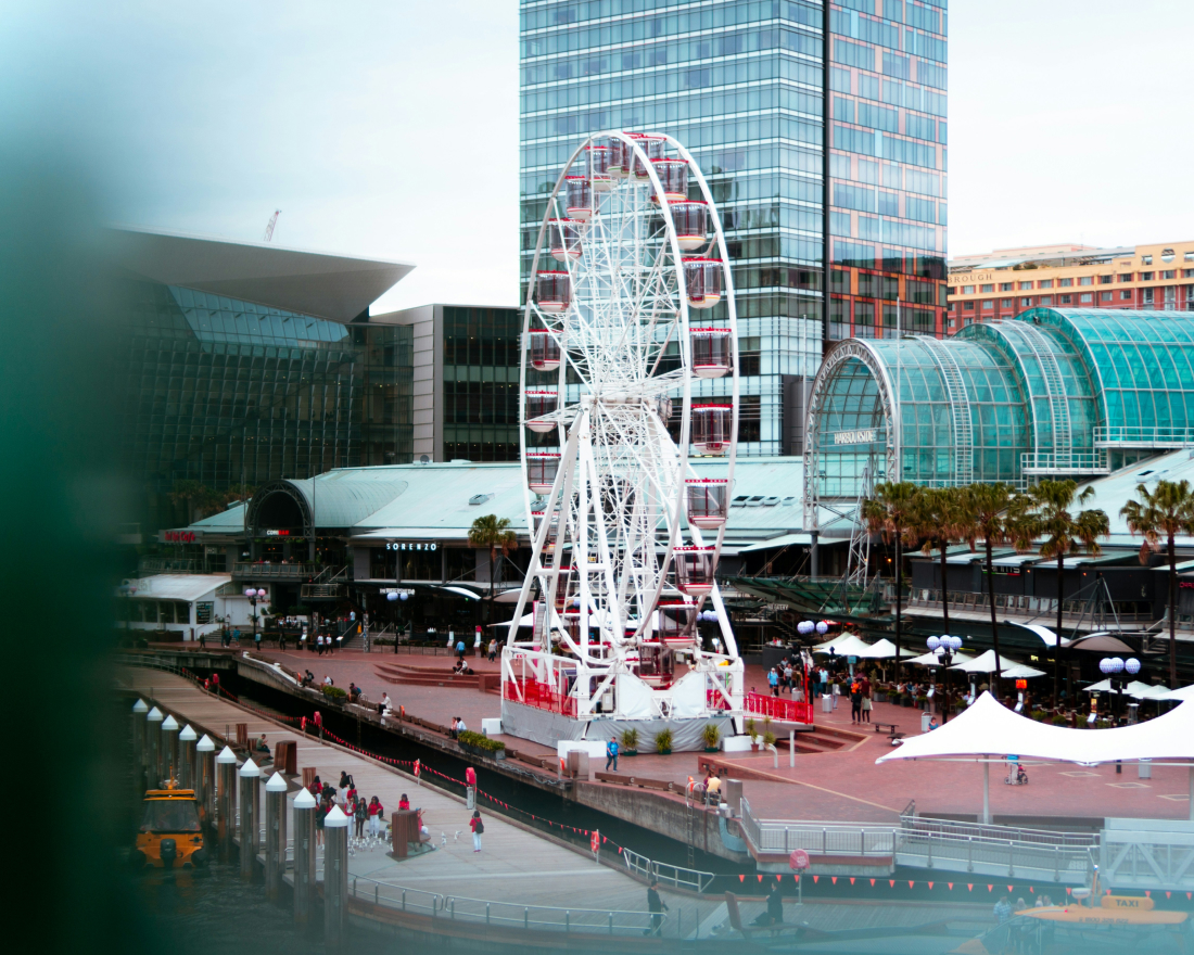 A vibrant urban waterfront scene featuring a large white ferris wheel, a bustling promenade with umbrellas, modern glass buildings, and a reflective body of water.