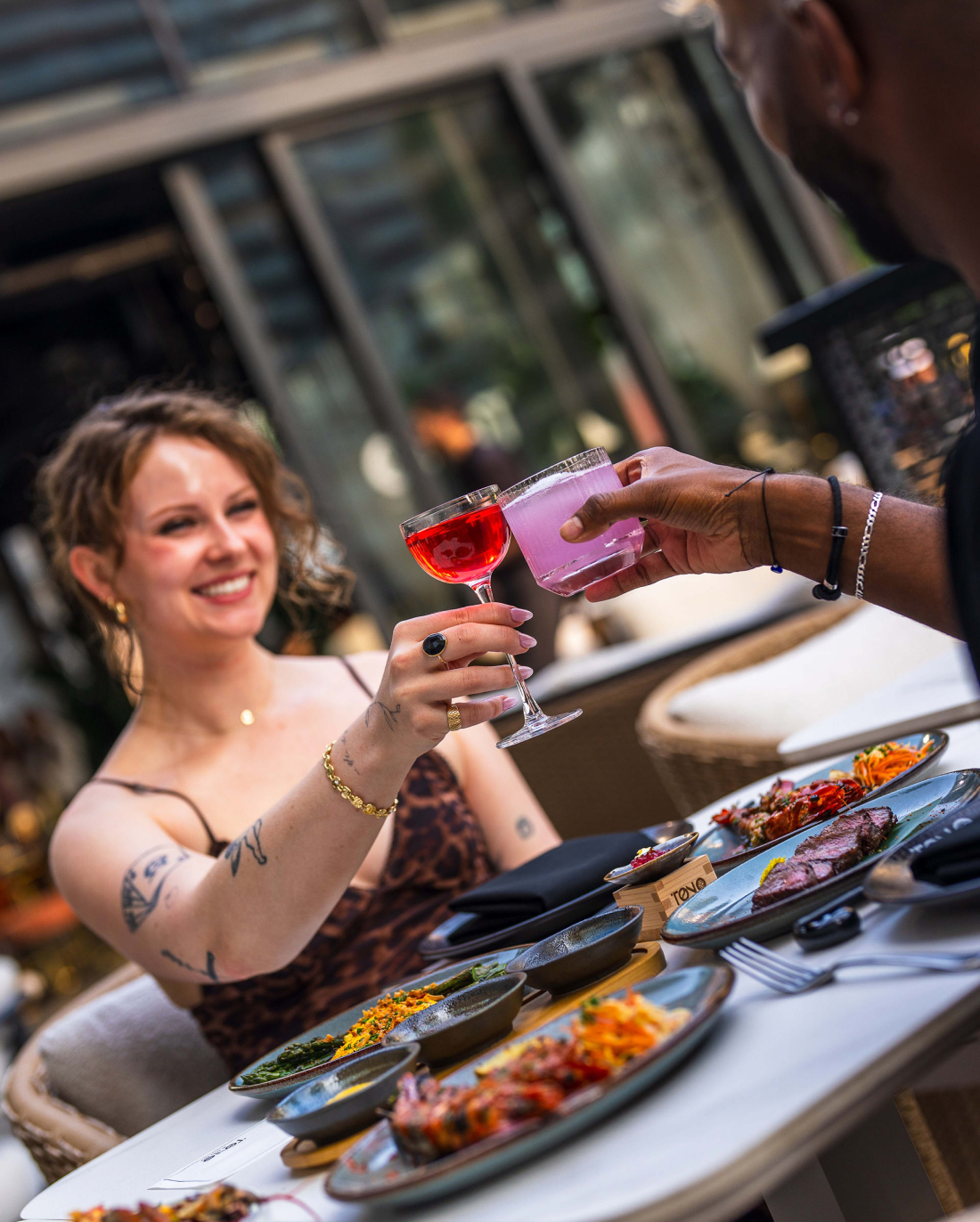 Dans un restaurant en terrasse, deux personnes trinquent avec des cocktails colorés devant une table garnie d'un assortiment de plats. L'une d'elles, vêtue d'une robe à imprimé léopard, sourit. L'ambiance semble décontractée et accueillante.