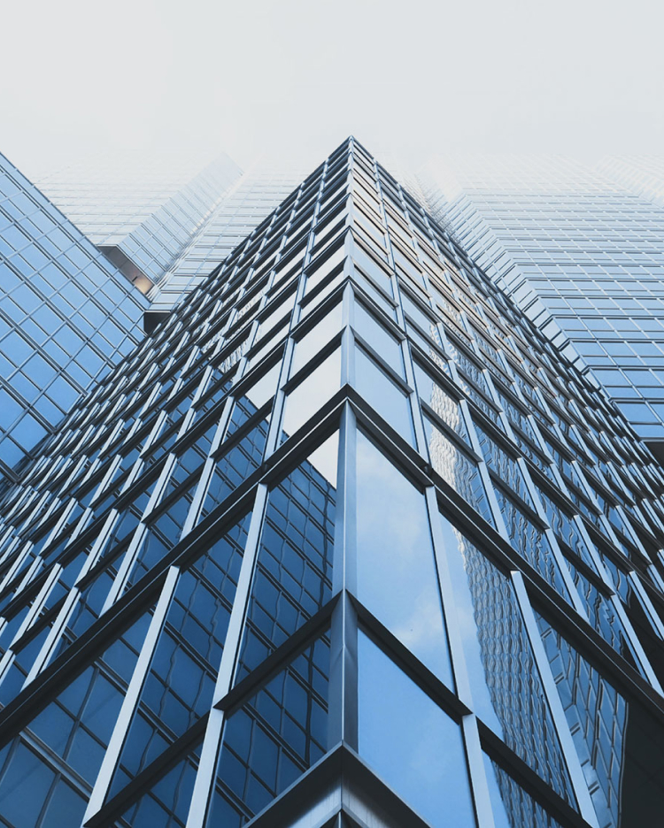 A low-angle view of a modern glass skyscraper, showing a series of reflective windows that intersect with metal beams, under a clear blue sky with clouds reflected on the surface.