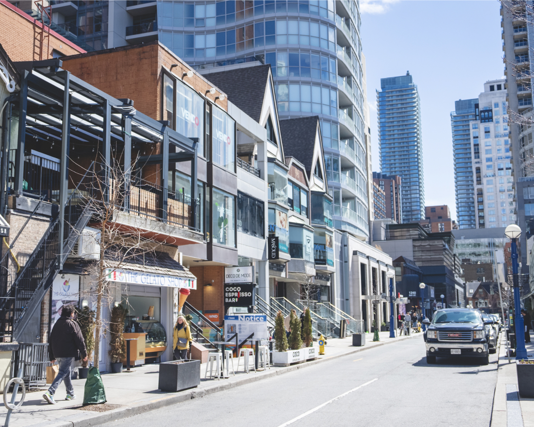 City street scene with modern and traditional buildings, including shops and restaurants at street level, under a clear blue sky. pedestrians and parked vehicles are visible.