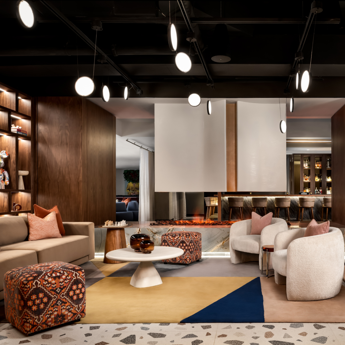 Salon moderne avec des canapés beiges, des poufs à motifs, une table basse ronde, un tapis géométrique et une cheminée centrale. L'éclairage chaleureux et les murs lambrissés créent une ambiance cosy et élégante. On aperçoit des étagères et un coin bar à l'arrière-plan.