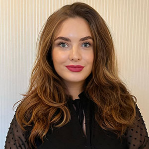 A woman with long, wavy brown hair wearing a black top stands in front of a light-colored, striped background. She has bright eyes, wears red lipstick, and is smiling softly at the camera.