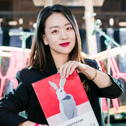 A woman with long, dark hair and wearing a black blazer holds up a framed picture of a white rabbit against a red background. She is smiling gently, standing in front of a blurred background with various colorful elements.
