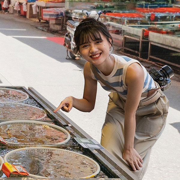 Una mujer sonriente, vestida con una camiseta de rayas y pantalones beige, con una cámara colgada del hombro, se agacha y señala cestas llenas de agua que contienen criaturas marinas en un entorno de mercado.