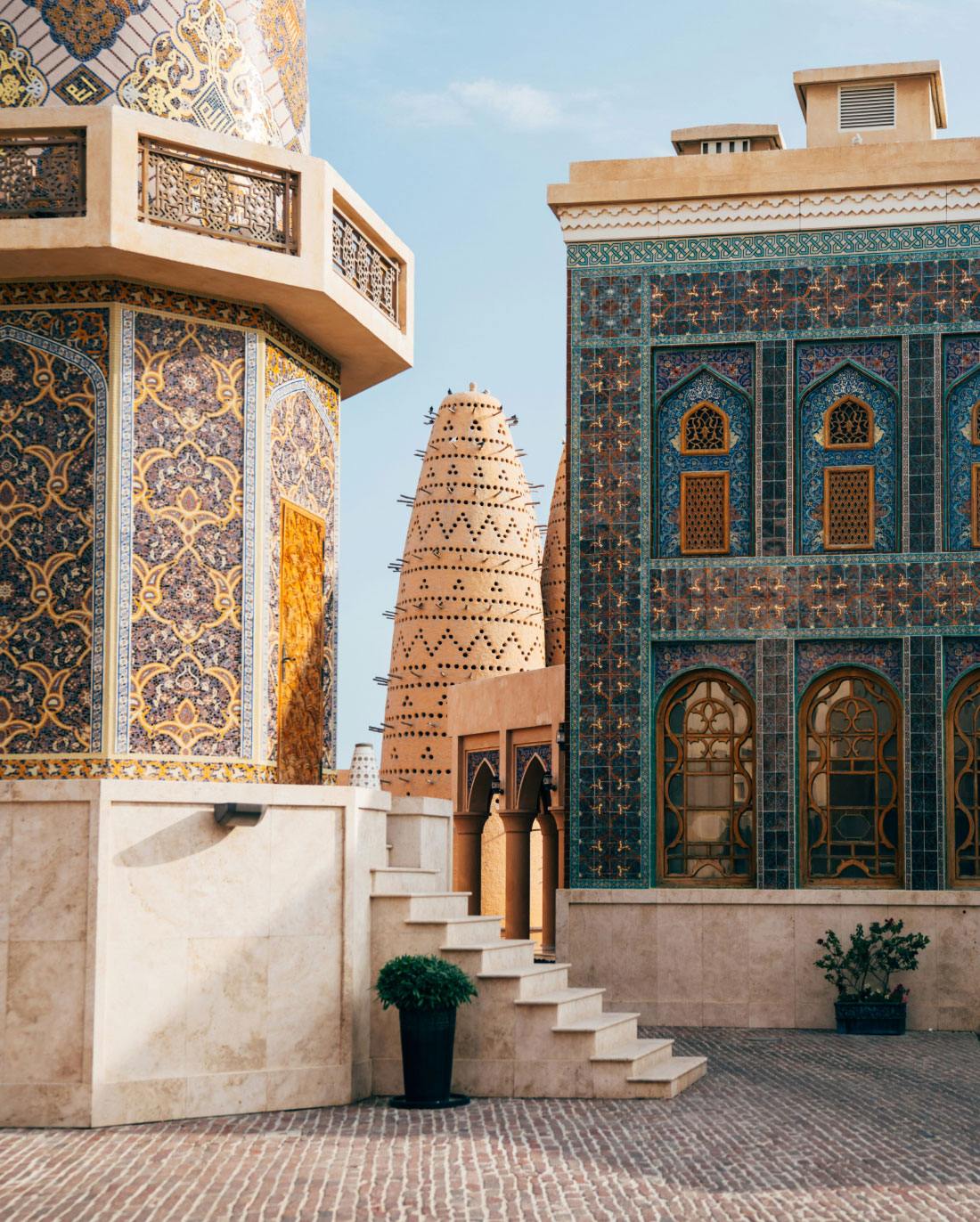 A picturesque courtyard features ornate buildings adorned with intricate tile work and colorful mosaics. Steps lead up to a wooden door, and a tall, conical structure with elaborate designs is visible in the background. Potted plants add a touch of greenery.
