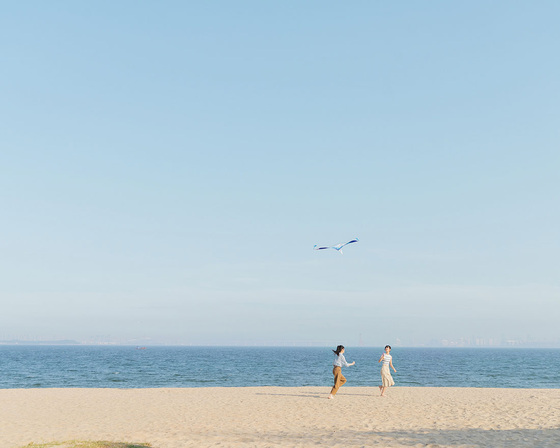 Dos personas hacen volar una cometa en una playa de arena con el océano de fondo. El cielo está despejado y azul y ambos visten ropa informal y ligera.