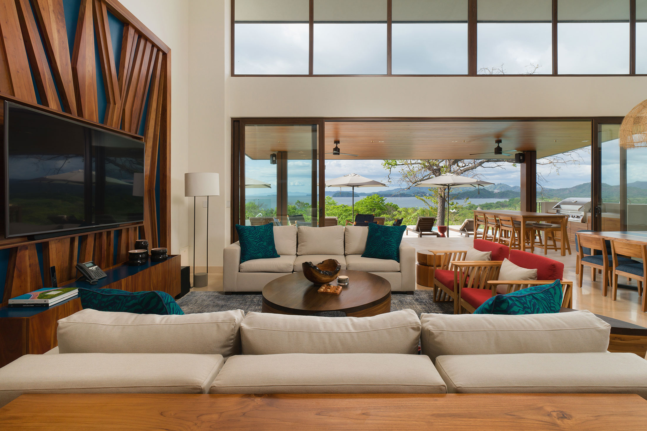 Un salon moderne et spacieux avec de grandes fenêtres offrant une vue panoramique sur un paysage verdoyant et luxuriant et des montagnes lointaines. La pièce est meublée de canapés beiges, d'une table basse en bois et d'une grande télévision. Un coin repas avec une table et des chaises en bois se trouve à proximité.