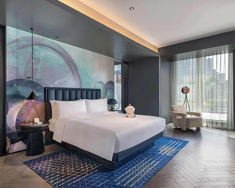 Habitación de hotel moderna con una cama grande, un cabecero oscuro y arte abstracto en la pared. Una alfombra geométrica azul se encuentra sobre un piso de madera. Hay una mesa auxiliar con una lámpara, un sillón cerca de grandes ventanales con cortinas transparentes y una vista del paisaje urbano desde el exterior.