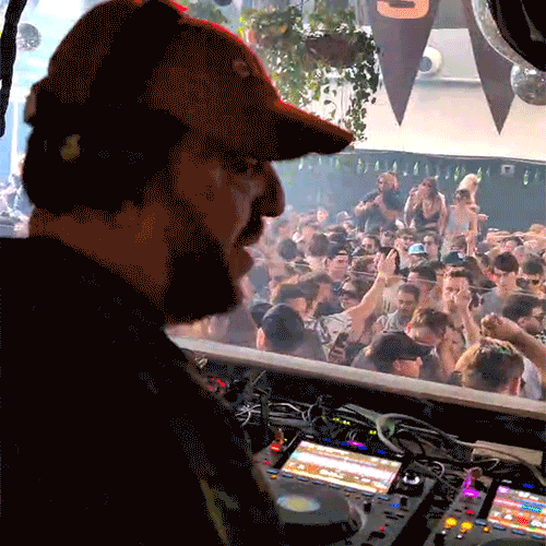 DJ mit roter Kappe und Kopfhörern spielt Musik in einem lebhaften Club. Die Menge ist voller Energie und tanzt unter bunten Lichtern und reflektierenden Discokugeln. Die Atmosphäre ist lebhaft und festlich.