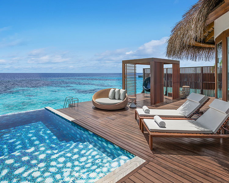 Luxuriöse Terrasse am Meer mit privatem Pool, zwei Liegestühlen mit weißen Handtüchern, einem runden gepolsterten Sitz und einer Cabana mit Strohdach und Blick auf das türkisfarbene Meer unter einem klaren blauen Himmel.