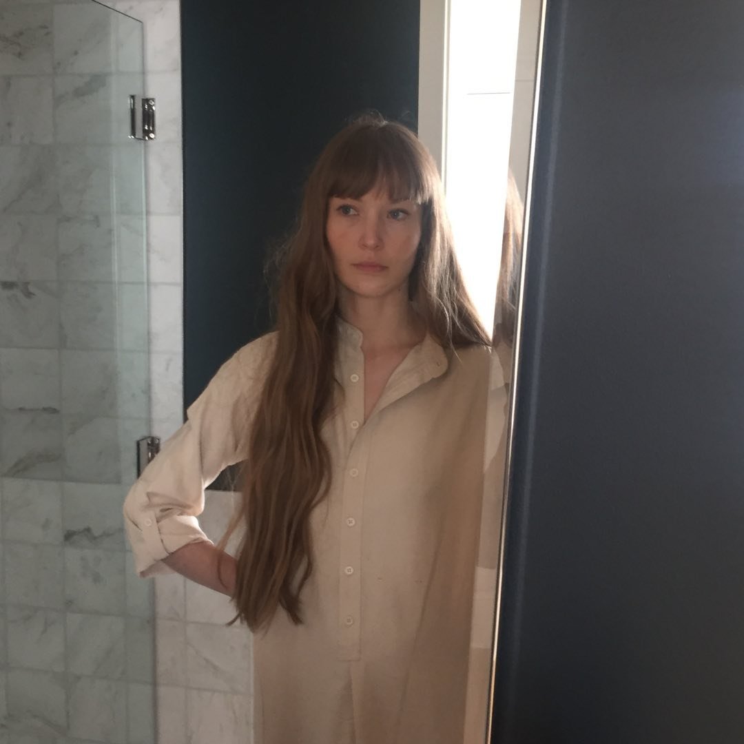 Una mujer de pelo largo y castaño se encuentra en un baño, con una camisa abotonada de color claro. Se mira en un espejo, con una ducha de azulejos visible al fondo. La iluminación es suave y natural.