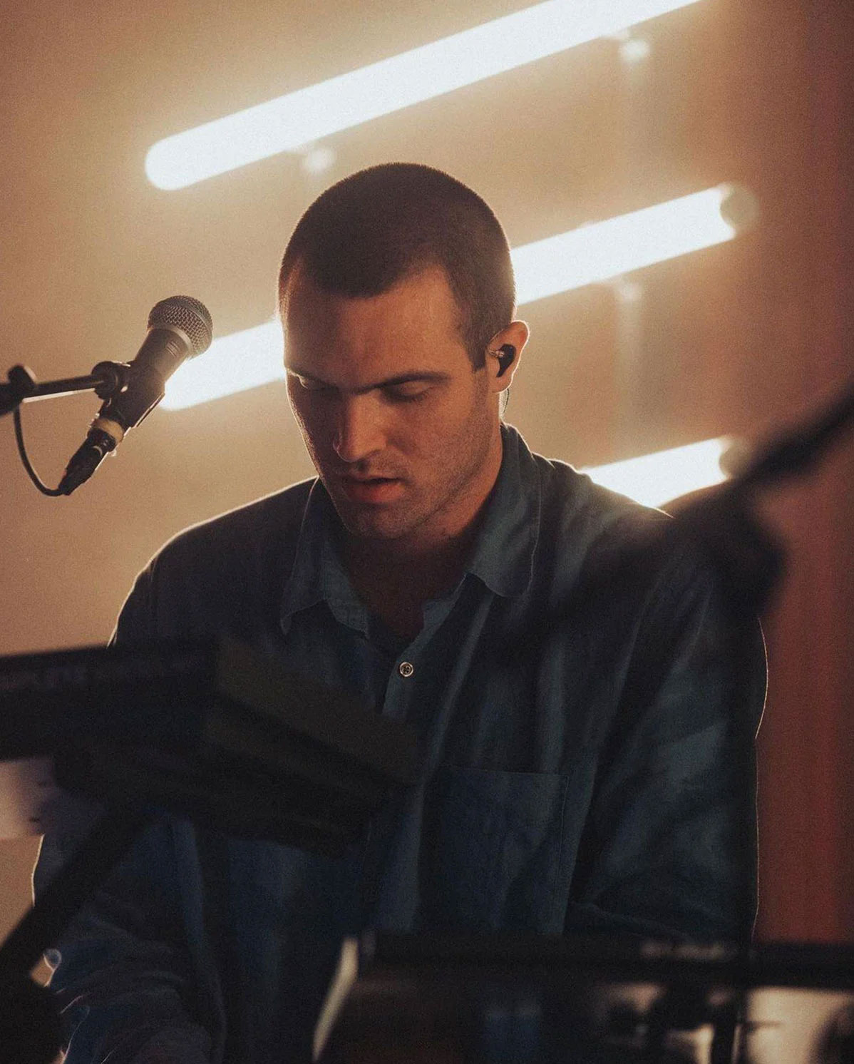 Ein Musiker spielt Keyboard und singt in ein Mikrofon auf der Bühne. Die Beleuchtung ist warm mit hellen horizontalen Lichtern im Hintergrund, was eine weiche, konzentrierte Atmosphäre schafft. Der Musiker trägt ein dunkles Hemd und scheint sich auf seinen Auftritt zu konzentrieren.