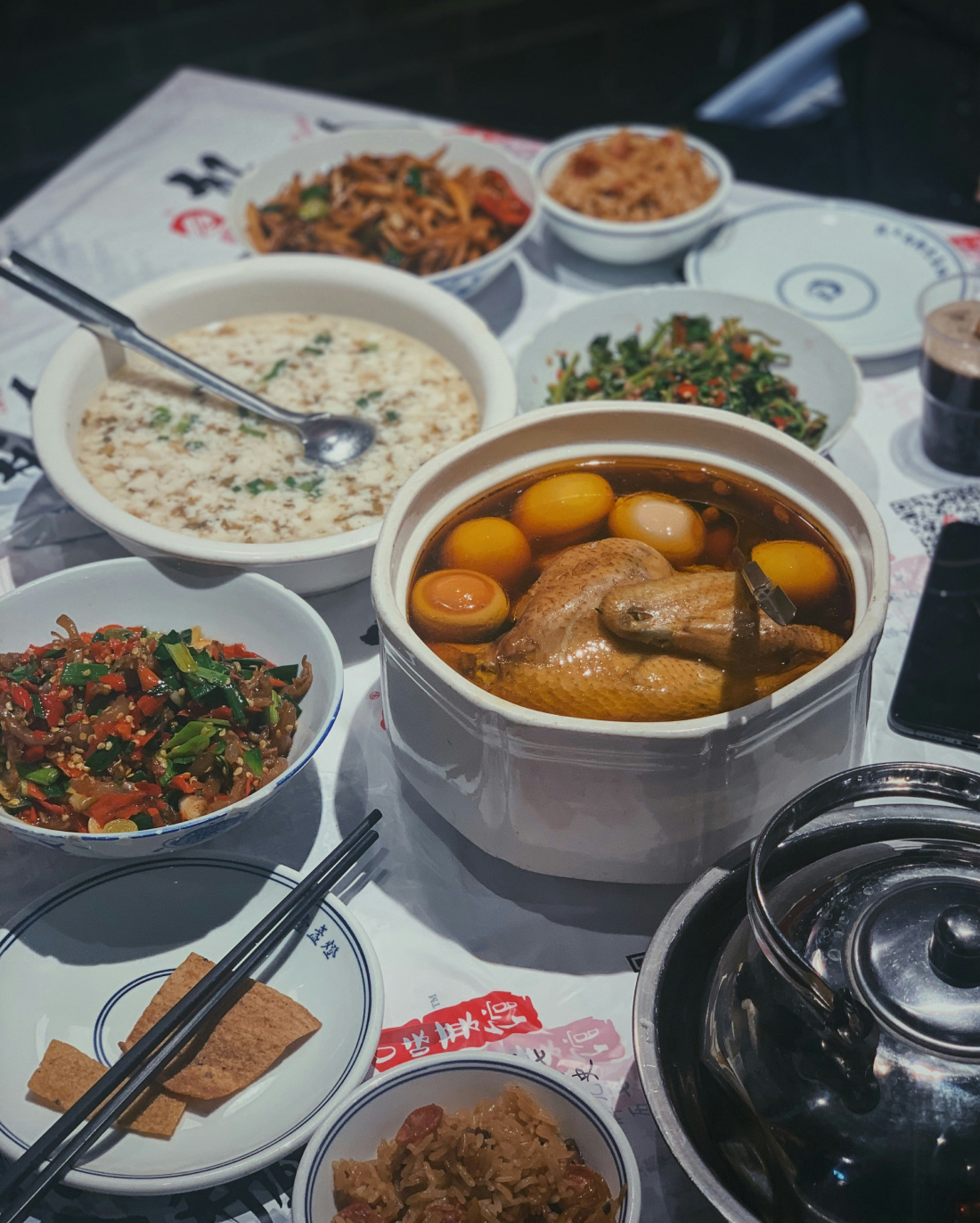 Una mesa repleta de platos chinos, entre ellos un pollo entero en caldo, gachas de arroz, verduras salteadas y un plato de carne picante. Platos y palillos están dispuestos a disposición, listos para comer.