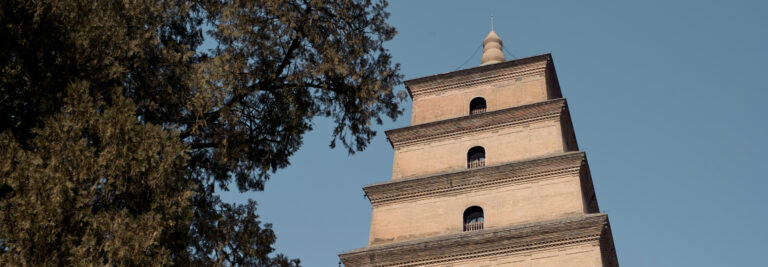 Esta pagoda alta e histórica se alza contra un cielo azul claro, rodeada de árboles frondosos. La estructura tiene varios niveles con aleros salientes y una parte superior puntiaguda. El ladrillo y la arquitectura tradicional reflejan un diseño antiguo.