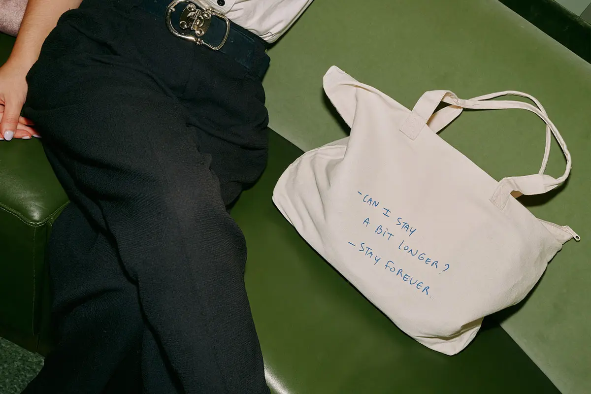 Se ve parcialmente a una persona con pantalones negros sentada en un sofá verde. A su lado hay una bolsa de mano beige con un texto que dice: 