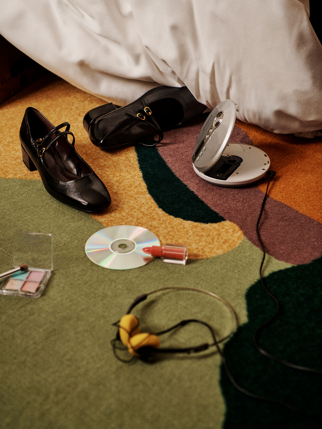 模様のあるカーペットの上に、黒いハイヒール、ポータブル CD プレーヤー、CD、口紅、アイシャドウ、黄色のヘッドフォンが置かれています。背景には白い布地の一部が見えます。.