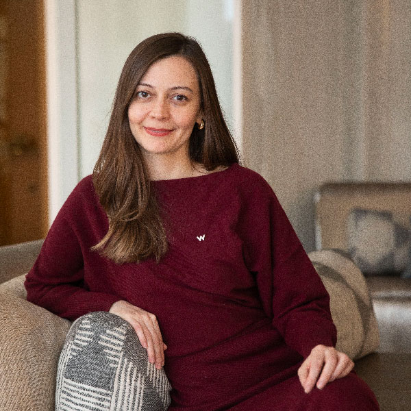 Una persona de cabello castaño largo está sentada en un sofá, con un suéter granate y sonriendo. La habitación tiene una decoración tranquila y neutra, con un cojín con un patrón geométrico.