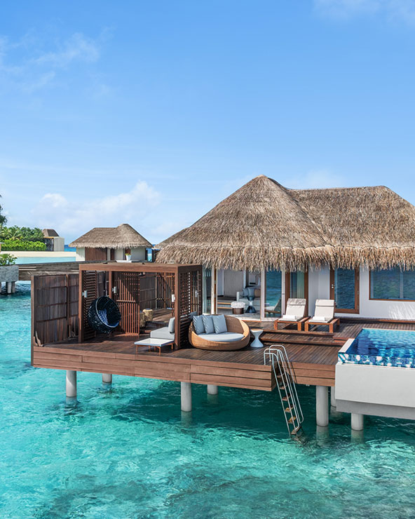 Ein luxuriöser Überwasserbungalow mit Strohdach thront über klarem, blauem Wasser. Auf der Terrasse stehen Liegestühle, ein Hängesessel und ein kleiner Pool mit einer Leiter, die zum Meer hinunterführt. Der Himmel ist hell und sonnig.