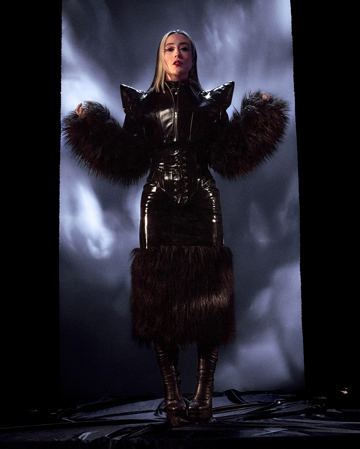 Eine Person steht selbstbewusst in einem dramatischen schwarzen Outfit mit glänzenden, strukturierten Schultern und großen, dunklen Pelzakzenten an Ärmeln und Rock vor einem stimmungsvollen, schattigen Hintergrund.