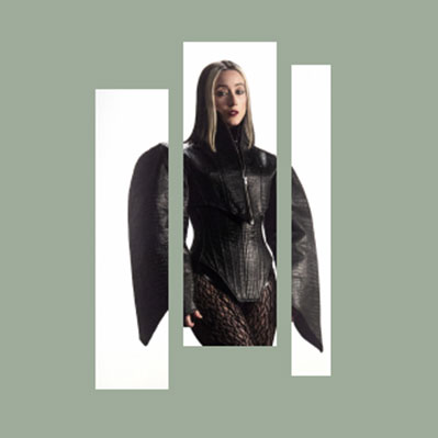 A person wearing a dramatic black outfit with exaggerated shoulders stands against a plain white background, framed by three vertical rectangular cutouts on a muted green backdrop.