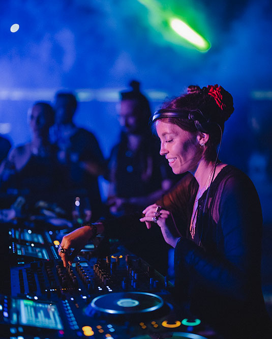 A DJ with headphones around her neck mixes music on a soundboard at a dimly lit club with blue and purple lighting and a green light beam in the background. People are watching her perform.