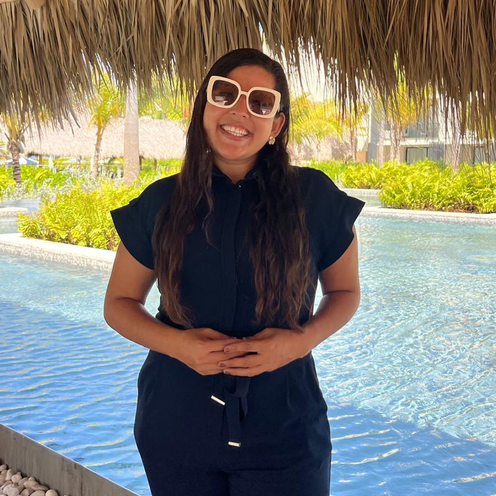 A woman with long hair and large white sunglasses smiles while standing in front of a pool, under a thatched roof, with tropical plants and palm trees in the background.