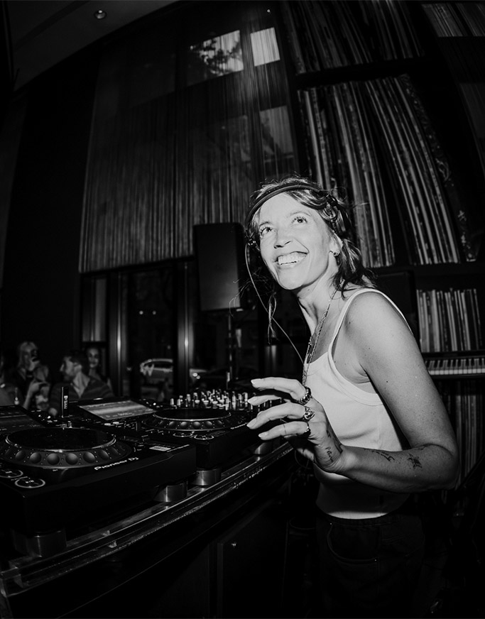 Eine lächelnde Frau mit Kopfhörern steht hinter einem DJ-Controller in einem schwach beleuchteten Raum mit hohen Regalen voller Schallplatten im Hintergrund. Sie scheint Musik zu mixen und den Moment zu genießen. Das Bild ist in schwarz-weiß gehalten.