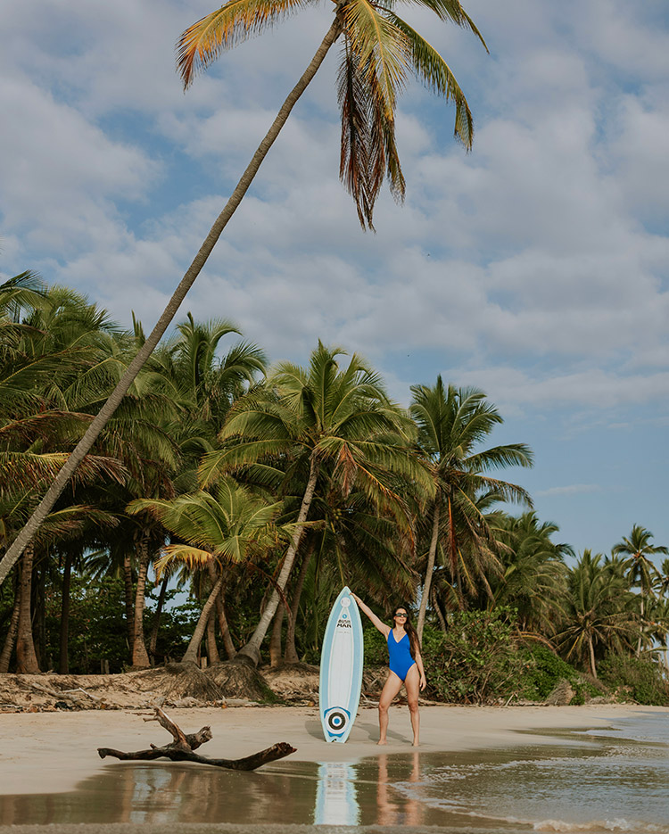 A woman in a blue swimsuit stands on a sandy beach holding a white surfboard, surrounded by tall palm trees under a partly cloudy sky.