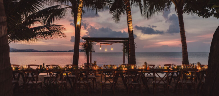 Una larga mesa de comedor al aire libre, decorada con velas y elementos tropicales, se encuentra en la playa bajo palmeras al atardecer, con vistas a un mar en calma y un cielo colorido de fondo.