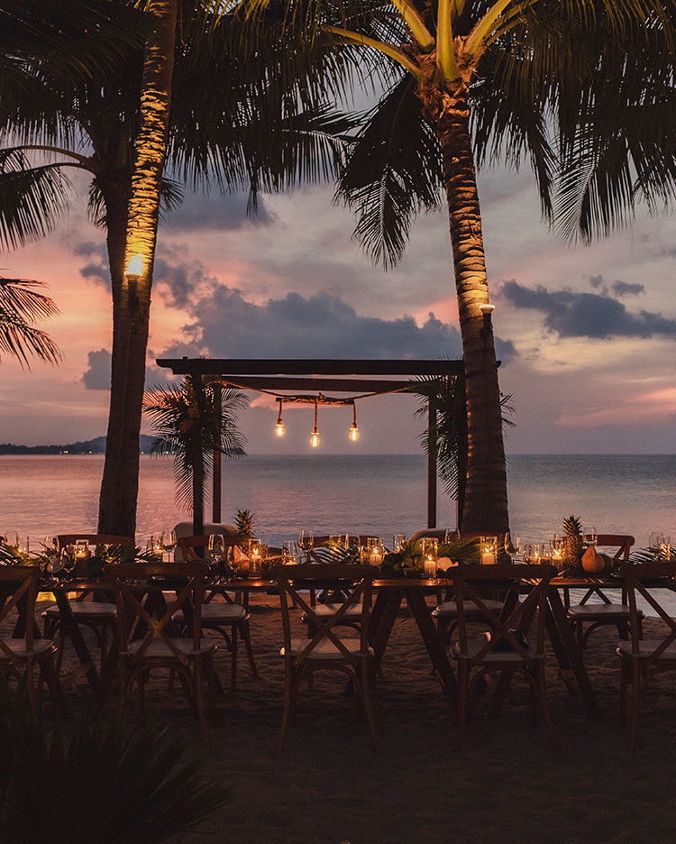 Ein langer Esstisch ist für das Abendessen bei Sonnenuntergang am Strand zwischen Palmen gedeckt, mit warmen Hängelampen, Kerzen und einem Blick auf das Meer und den farbenfrohen Himmel im Hintergrund.