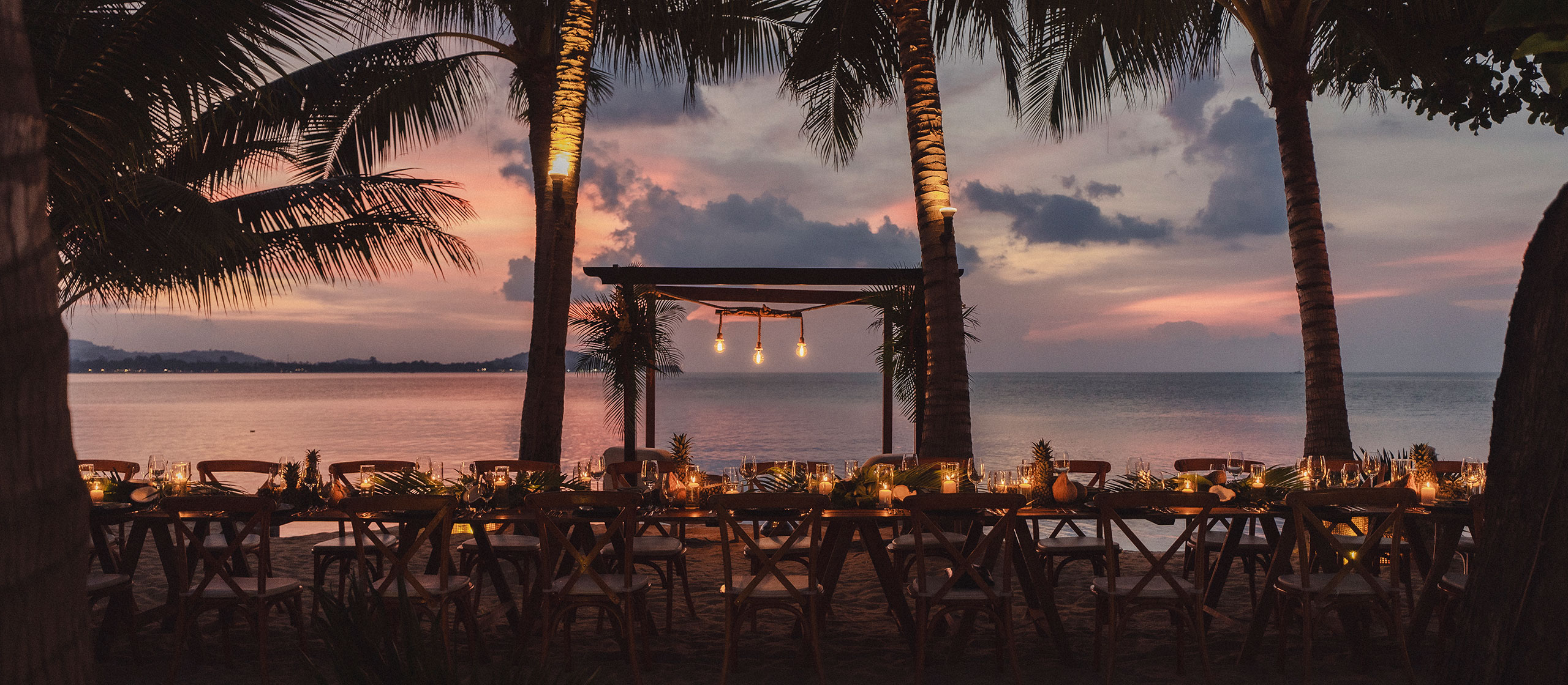 Lange Esstische im Freien mit Kerzen und Dekorationen stehen bei Sonnenuntergang an einem Sandstrand unter Palmen und bieten Aussicht auf das ruhige Meer und einen teilweise bewölkten Himmel mit rosa und violetten Farbtönen.
