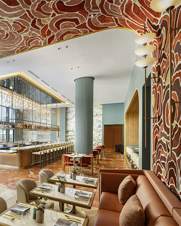 Ein modernes Restaurant mit abstrakt gemusterter Decke und Wänden in kräftigem Rot und Beige, eleganter Barbestuhlung, hellgrünen Säulen, Marmorböden und stilvoll mit Tellern und Gläsern gedeckten Tischen.