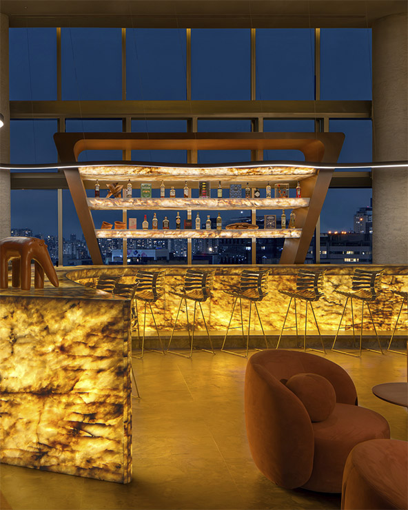 Eine moderne Bar bei Nacht mit leuchtenden, bernsteinfarbenen Theken und Regalen, eleganten Barhockern, braunen Loungesesseln und großen Fenstern mit der Skyline der Stadt im Hintergrund.