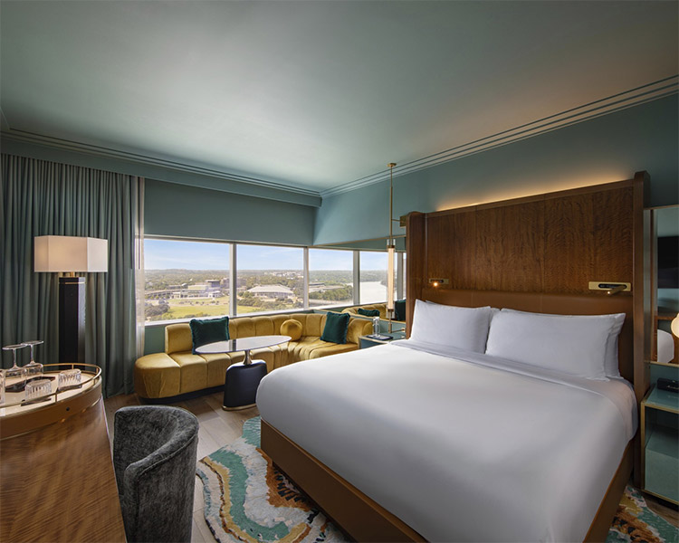 Modernes Hotelzimmer mit großem Bett, hölzernem Kopfteil, gelbem Sofa und großen Fenstern mit Panoramablick. Zur Einrichtung gehören eine sanfte Beleuchtung, ein runder Teppich und elegante Möbel in neutralen und pastellfarbenen Tönen.