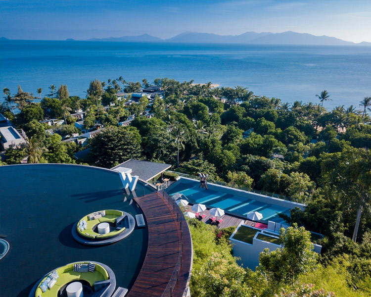 Luftaufnahme eines üppigen tropischen Resorts mit einer Lounge auf dem Dach, einem Pool mit Sonnenliegen und Palmen, mit Blick auf das blaue Meer und die fernen Berge unter einem klaren Himmel.