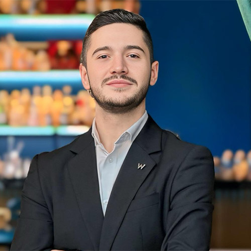 Ein junger Mann in einem dunklen Anzug und hellem Hemd steht selbstbewusst mit verschränkten Armen da und lächelt. Hinter ihm stehen Regale mit verschwommenen Flaschen und eine blaue Wand, die eine professionelle, moderne Atmosphäre schaffen.