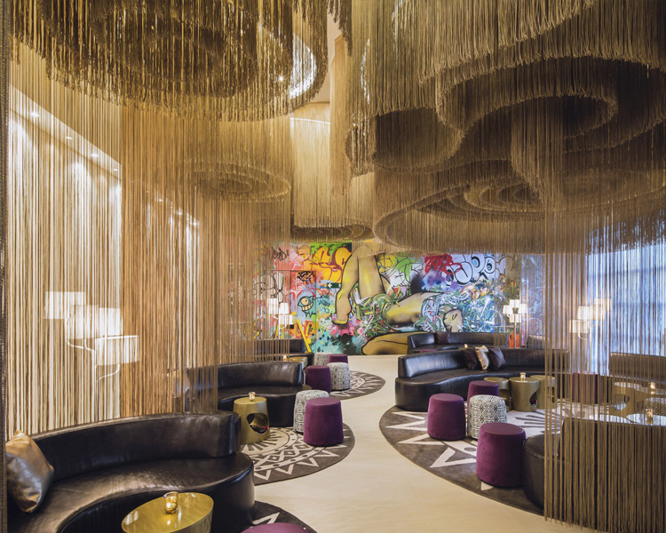 Eine stilvolle Lounge mit runden schwarzen Ledersofas, lilafarbenen Hockern, goldenen Tischen und kunstvoll aufgehängter Schnurdekoration. Die Rückwand zeigt bunte Graffiti-Kunst mit einer liegenden Figur und lebhaften Mustern.