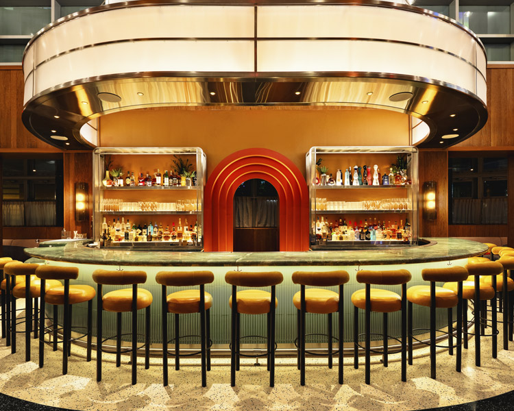 Eine stilvolle Bar im Retro-Stil mit einem geschwungenen Tresen, der von gelben, gepolsterten Hockern gesäumt wird, beleuchteten Flaschenregalen und einem roten Bogen in der Mitte, eingebettet in eine warme, moderne Einrichtung.