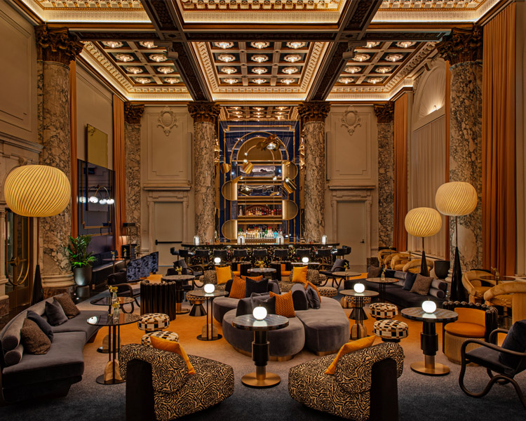 Eine luxuriöse Hotellounge mit verschnörkelten Marmorsäulen, hohen Kassettendecken, eleganter Beleuchtung, plüschigen Sitzgelegenheiten in dunklen Tönen, gemusterten Stühlen und einer stilvollen Bar im hinteren Bereich, die eine warme, elegante Atmosphäre schafft.