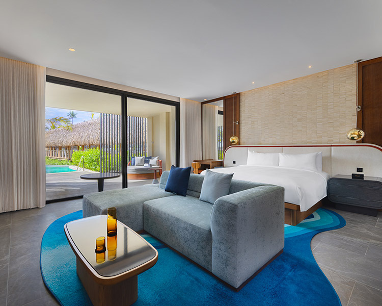 Modernes Hotelzimmer mit einem großen Bett, einem grauen Sofa, einem Couchtisch mit bernsteinfarbenen Gläsern, bodentiefen Fenstern und Blick auf einen privaten Innenhof mit grünem Gras und einem Pool draußen. Die Einrichtung ist schlicht und einladend.