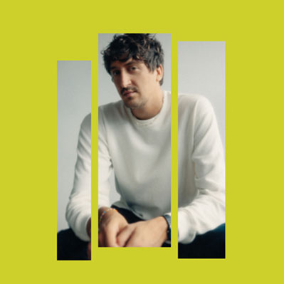 A man with curly hair and a mustache, wearing a white sweater, sits against a light background. The image is divided into three vertical panels set on a solid yellow-green background.