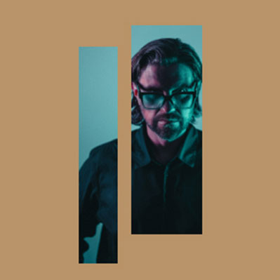 A man with glasses and shoulder-length hair is lit with blue and green light, viewed through two vertical rectangular cutouts on a tan background.