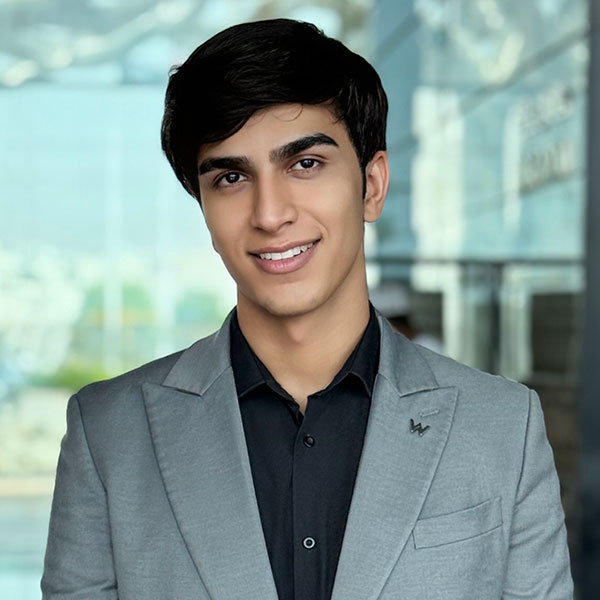 Un joven moreno con traje gris claro y camisa negra sonríe a la cámara. El fondo es moderno y está suavemente difuminado.
