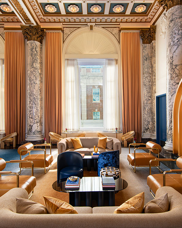 Elegantes Wohnzimmer mit Marmorsäulen, hoher verzierter Decke, großen Fenstern mit goldenen Vorhängen, modernen beigen Sofas, goldenen Sesseln und einem zentralen Couchtisch mit Büchern, alles in natürliches Licht getaucht.