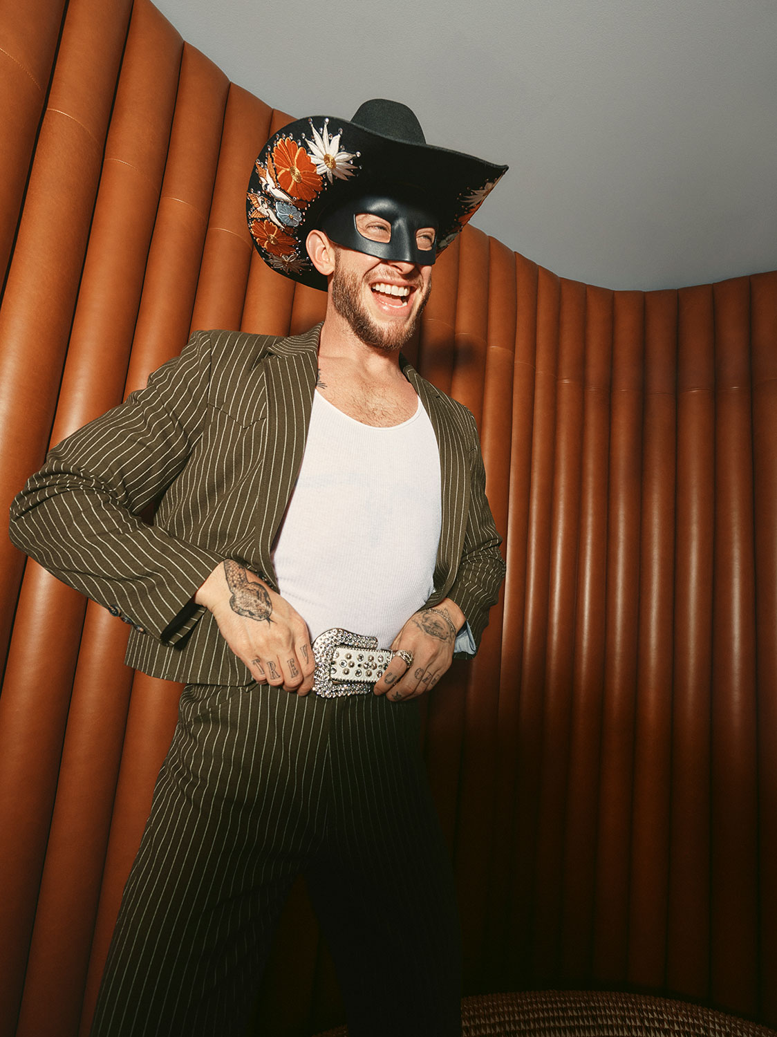 A smiling man wearing a pinstriped suit, white tank top, black cowboy hat with floral decorations, and a black mask stands in front of a brown, padded wall. He is holding his belt with both hands.