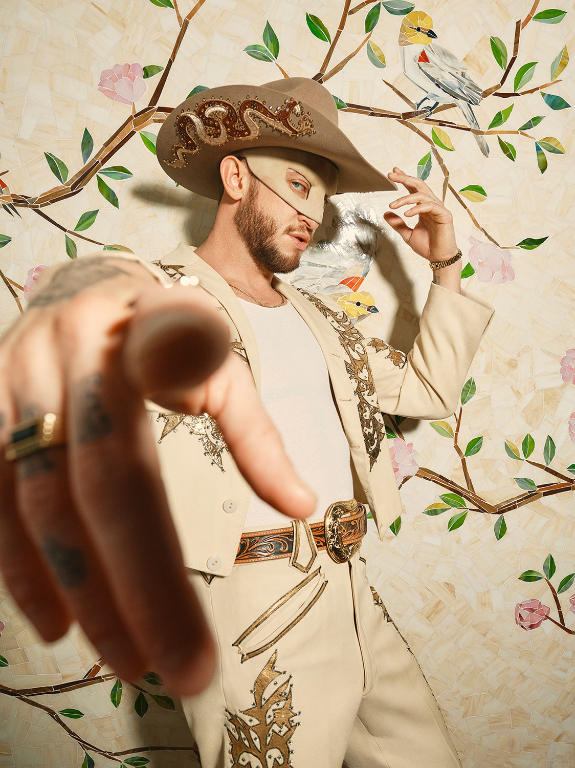 A person in an ornate beige suit and wide-brimmed hat with a snake design points toward the camera. They stand in front of a decorative wall featuring painted tree branches and birds.