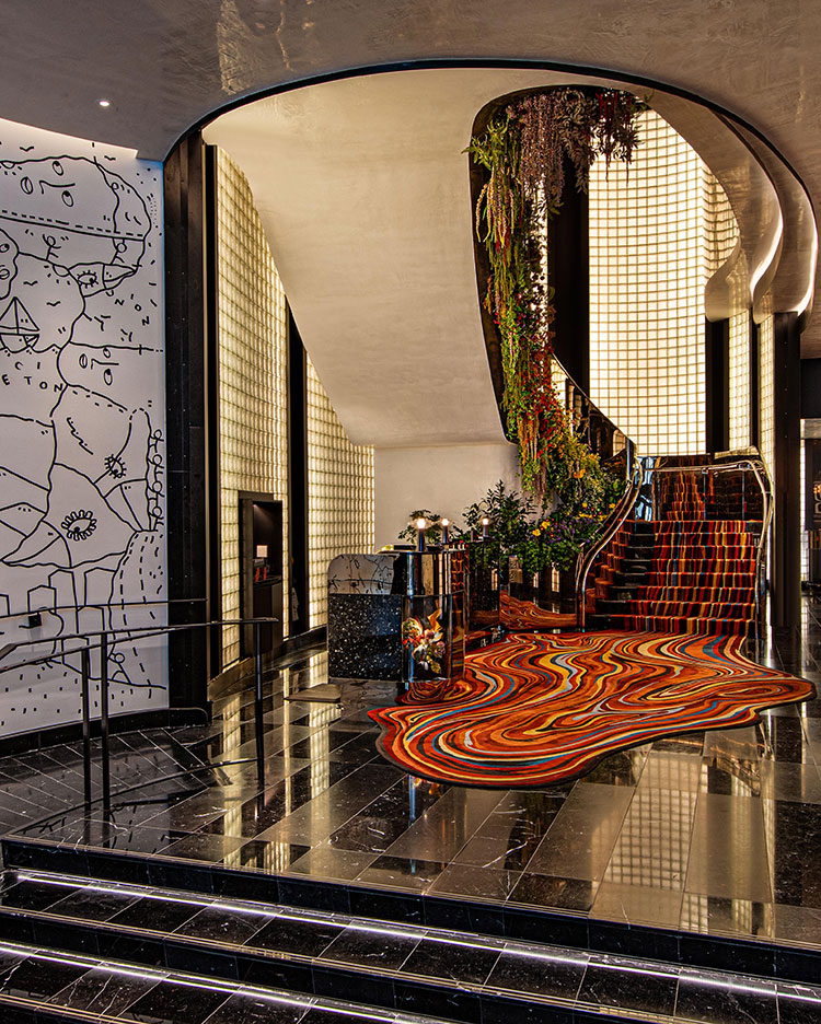 Eine moderne Hotellobby mit glänzenden schwarzen Marmorböden, einem farbenfrohen gewellten Teppich, einer großen, mit Pflanzen geschmückten Treppe, Glasblockwänden und einer großen weißen Wand mit abstrakter schwarzer Linienkunst.