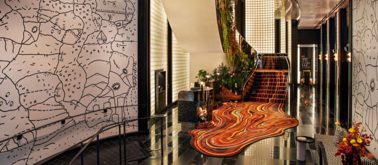 Un elegante vestíbulo de hotel con suelos negros brillantes, una vibrante alfombra ondulada de color rojo anaranjado, plantas en las escaleras y paredes blancas con arte lineal abstracto en negro que recuerda a un mapa. El espacio es moderno y tiene un diseño creativo.