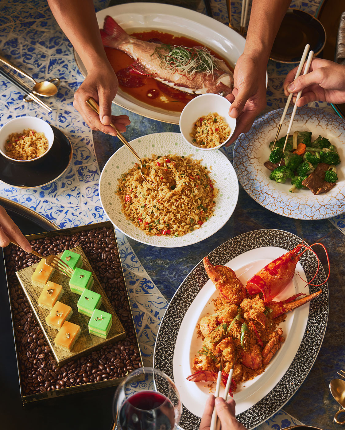 Una mesa puesta con varios platos asiáticos, entre ellos arroz frito, langosta, un pescado entero, verduras con ternera, postres, y varias manos usando palillos y cucharas de servir para compartir la comida.