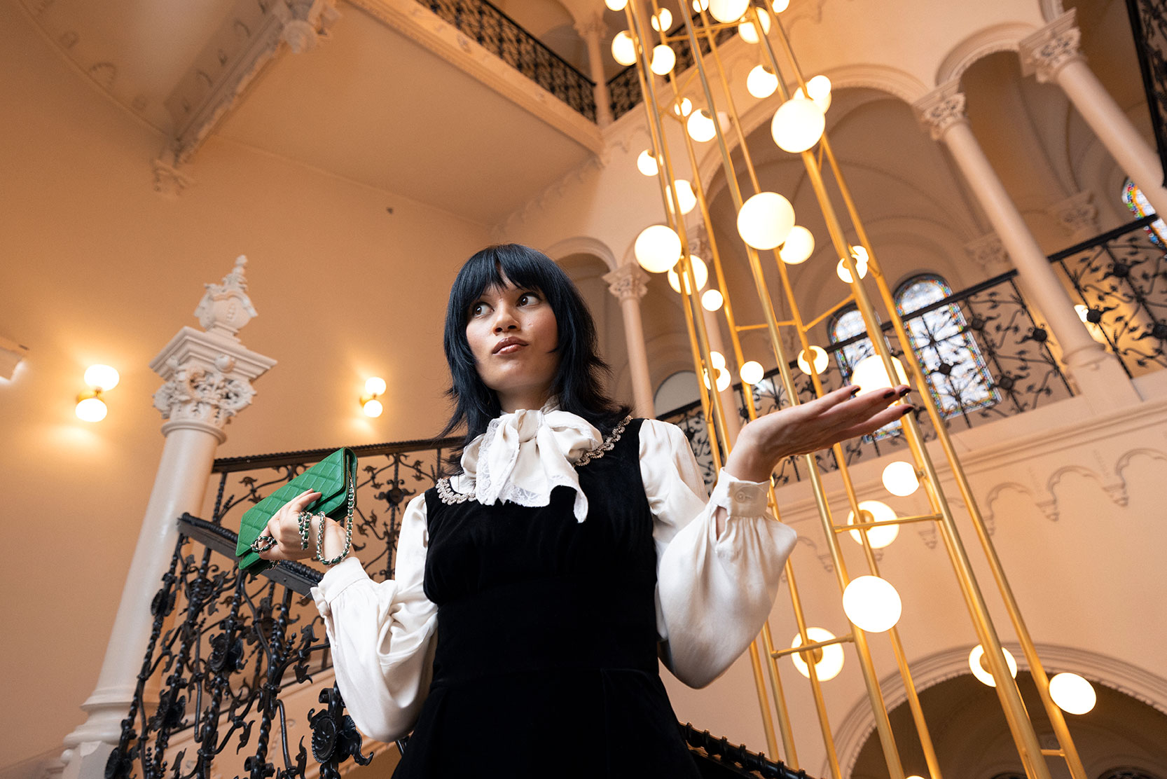 A woman in a black dress with a white ruffled blouse stands indoors, holding a green purse and gesturing with one hand. Ornate railings, arches, and a tall chandelier with glowing bulbs surround her.